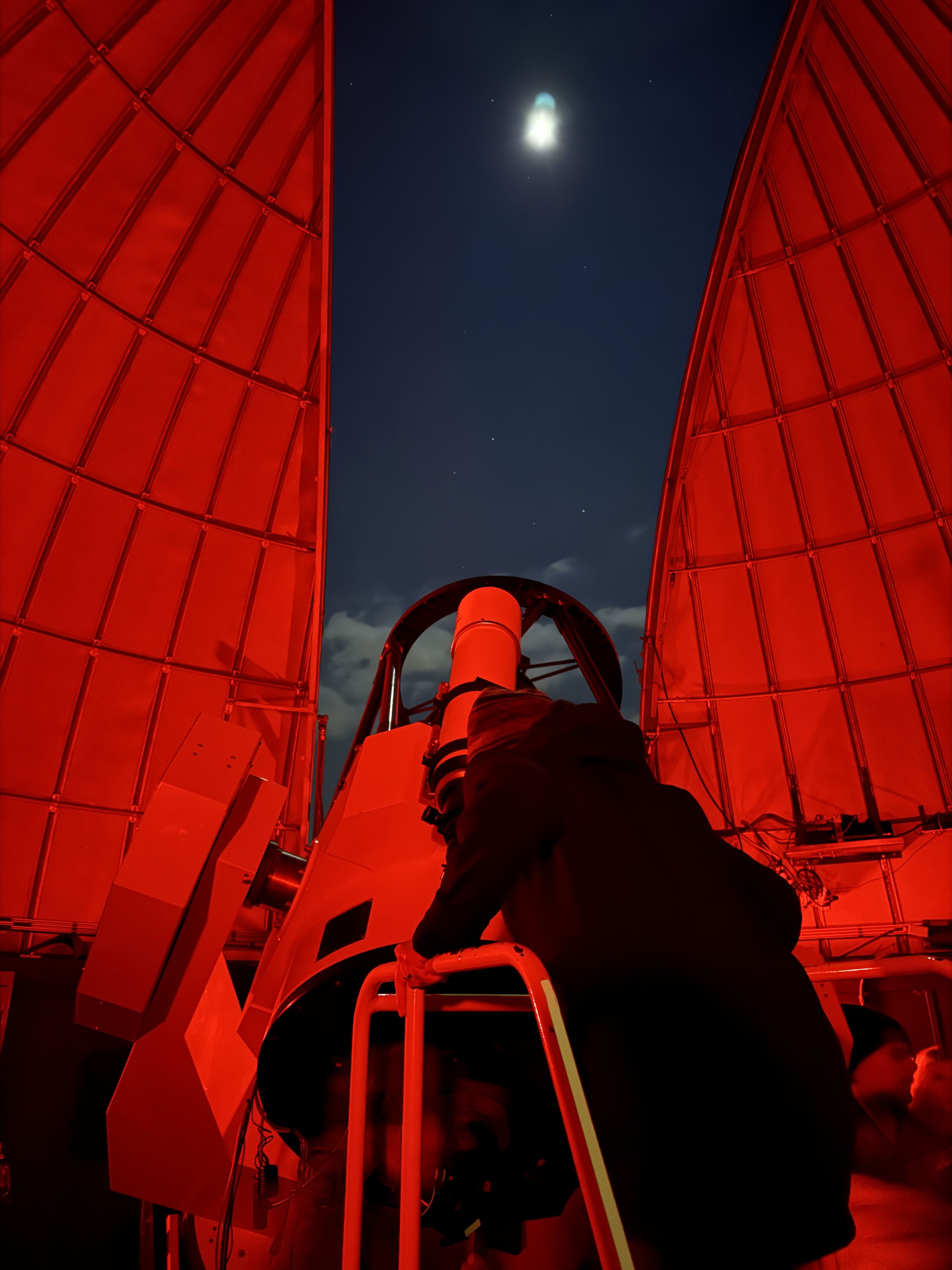 A wide-angle photograph captures a person using a large telescope inside an observatory dome at night. The image is bathed in red light, which preserves night vision for astronomical observations. Rebecca is positioned at the eyepiece of the Schulman 32-inch telescope at the University of Arizona's Mount Lemmon SkyCenter, her silhouette clearly defined against the illuminated equipment. The observatory's dome opening creates a natural frame that divides the composition, revealing a dark night sky with the moon visible as a bright white orb surrounded by scattered stars. The curved walls of the dome display a geometric pattern of panels, all tinted in the same red hue that dominates the scene. The telescope mount and mechanical components are clearly visible. The contrast between the warm red interior lighting and the cool darkness of the night sky beyond creates a clear separation between the controlled observatory environment and the vast cosmos being observed.