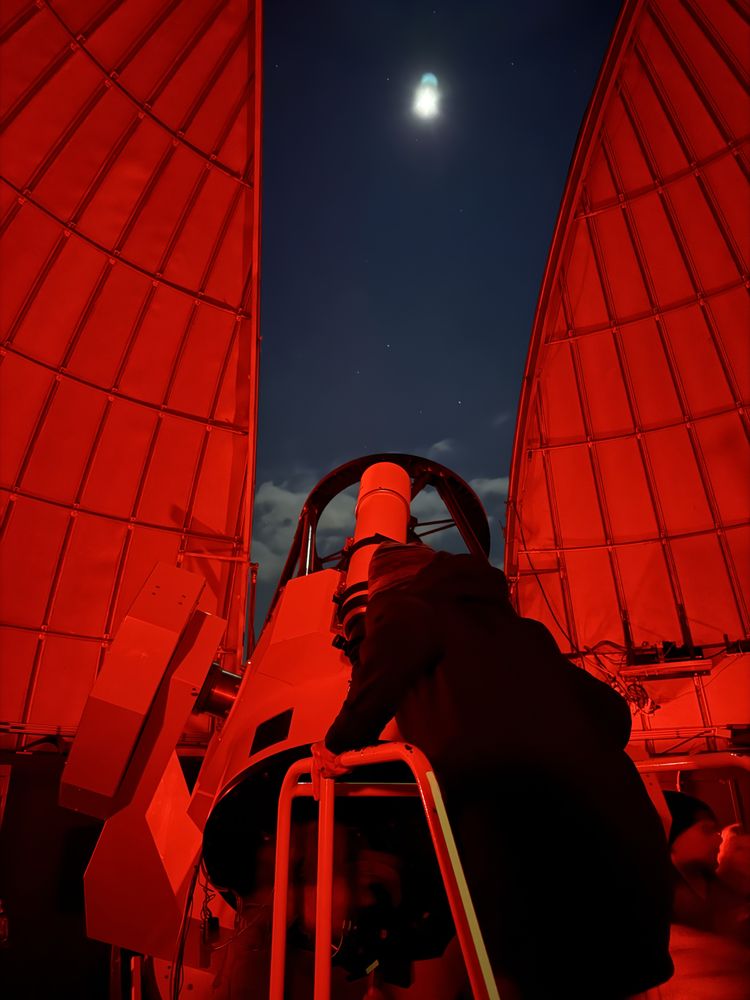 A wide-angle photograph captures a person using a large telescope inside an observatory dome at night. The image is bathed in red light, which preserves night vision for astronomical observations. Rebecca is positioned at the eyepiece of the Schulman 32-inch telescope at the University of Arizona's Mount Lemmon SkyCenter, her silhouette clearly defined against the illuminated equipment. The observatory's dome opening creates a natural frame that divides the composition, revealing a dark night sky with the moon visible as a bright white orb surrounded by scattered stars. The curved walls of the dome display a geometric pattern of panels, all tinted in the same red hue that dominates the scene. The telescope mount and mechanical components are clearly visible. The contrast between the warm red interior lighting and the cool darkness of the night sky beyond creates a clear separation between the controlled observatory environment and the vast cosmos being observed.