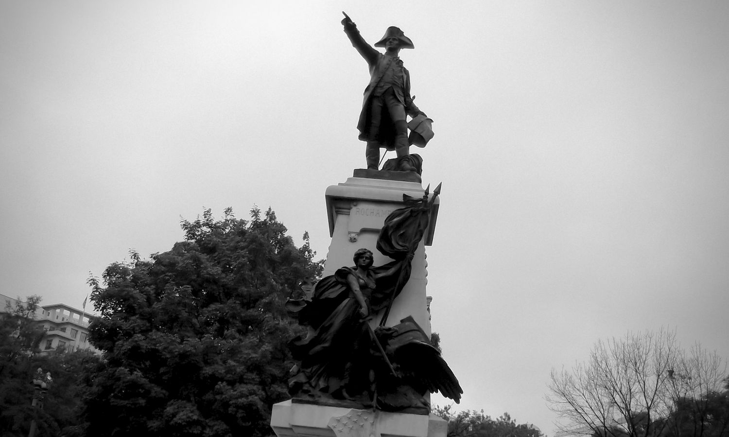 A tilted black and white photo of the statue Major General Comte Jean de Rochambeau in Lafayette Square in Washington, D.C., across the street from the White House. Rochambeau stands above Lady Liberty, pointing ahead.