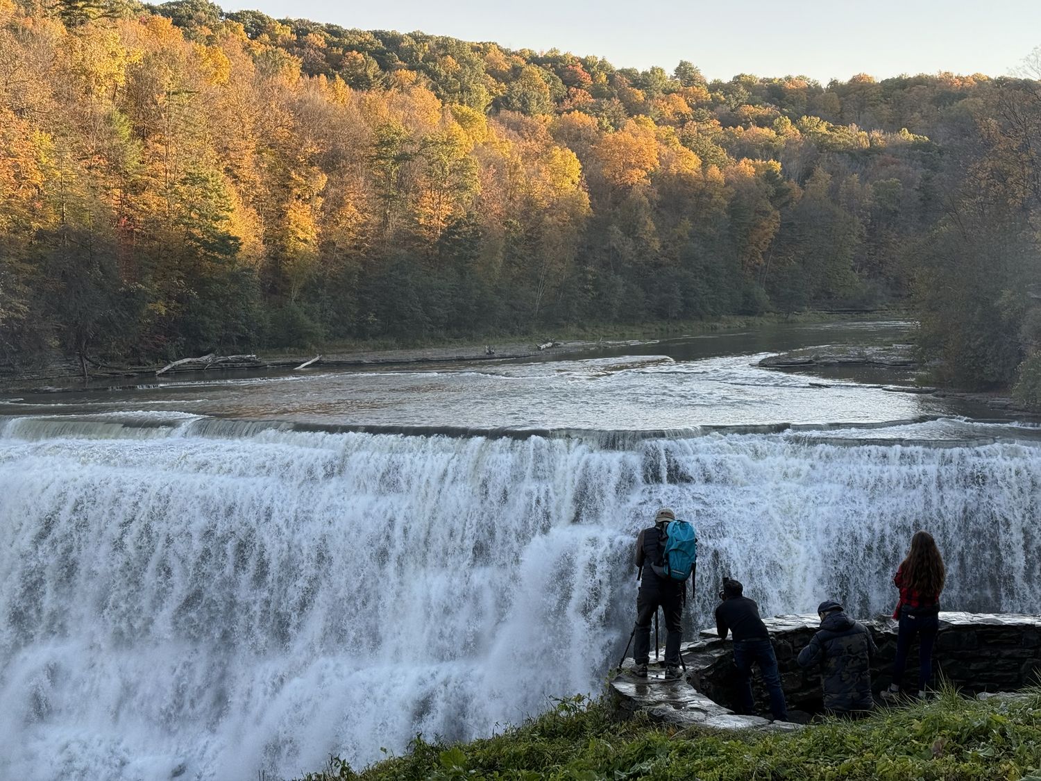 The Genesee River flows over the Middle Falls in Letchworth State Park. Trees on the bank of the river are turning to fall covers as the low sun illuminates them. In the foreground, several people stand on an overlook photographing the falls. One of them is somewhat precariously standing on a wet rock wall with a tripod.