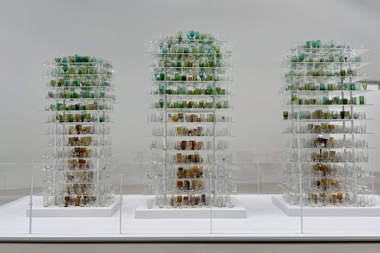 Three tall, tower-like sculptural columns made of hundreds of glass drinking vessels — goblets, tumblers, wine glasses, and cups — arranged on transparent acrylic shelves. Within each tower, the glasses are organized by color to form the shape of a tree: clear and amber-toned pieces at the base suggest a trunk, while green and teal glasses cluster toward the top to form a leafy crown. Each tower rises from a white pedestal behind a low glass barrier, set against a plain white gallery wall. The installation is “Forest Glass” by Katherine Gray.