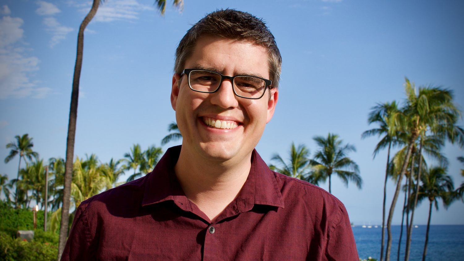 An outdoor portrait of Eric McCarthy, a white male. He’s looking at the camera and smiling while wearing glasses and a red collared button up T-shirt. Behind him are out-of-focus palm trees, a body of water, and what might be a few white sailboats.