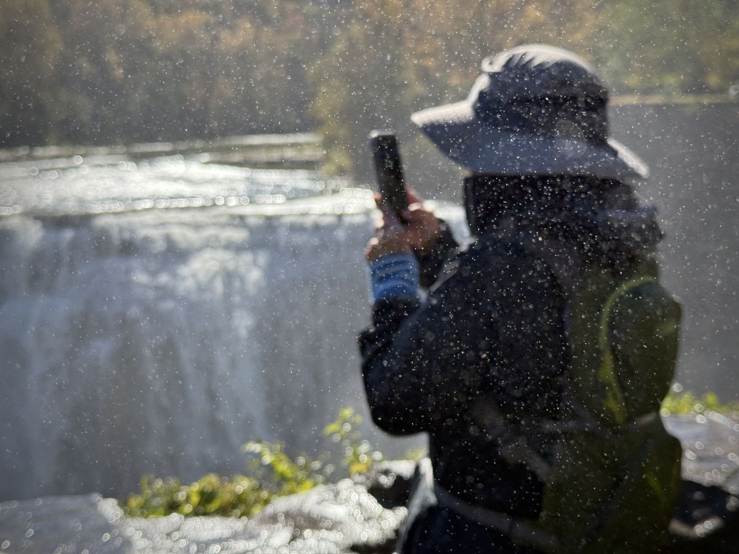 Sun-lit mist scatters in the foreground. A blurry person in a hat with a backpack uses their phone to take a photograph of a waterfall.