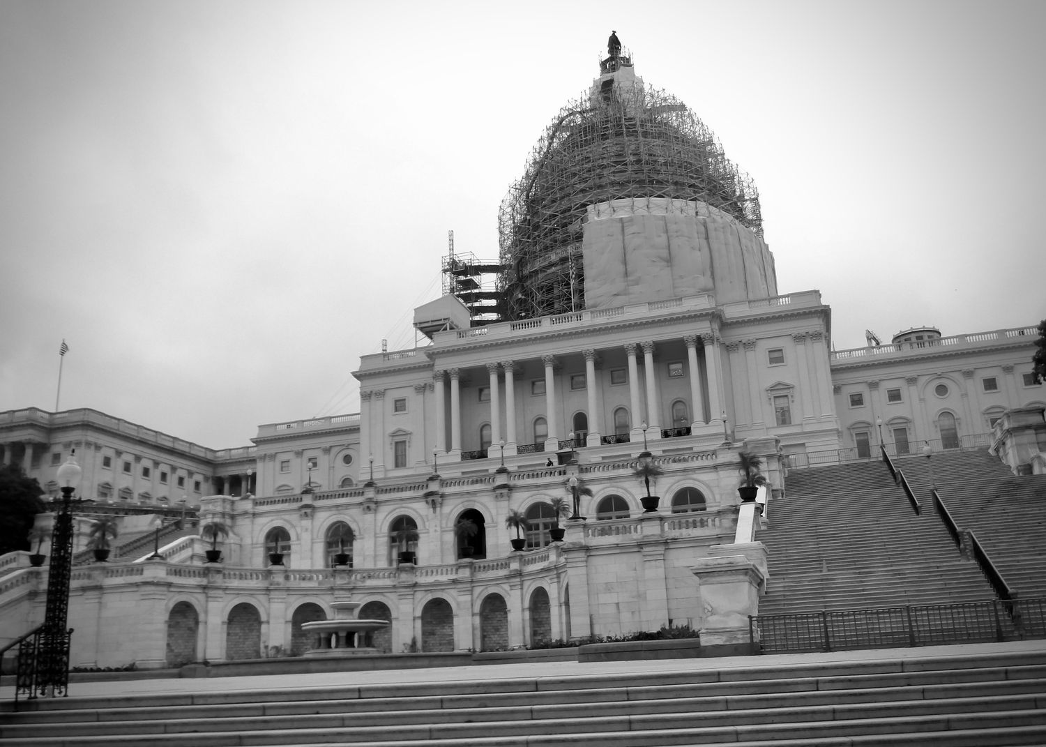 A slightly-askew black and white photo of the east side of the U.S. Capitol Building, taken from its lowest steps. The Capitol Dome is covered in scaffolding as it undergoes a restoration project.