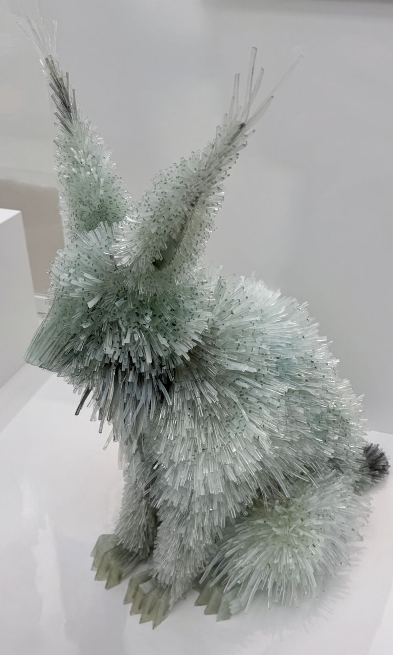 A sculpture of a seated lynx made entirely from thousands of small, pale green-tinted glass shards, giving the animal a spiky, crystalline fur texture. The lynx sits upright on a white pedestal with its tall, tufted ears prominently raised. The translucent glass catches the light, creating a shimmering, frost-like appearance against a plain white background. The work is “Lynx After a Sketchbook Page by Albrecht Dürer” by Marta Klonowska.