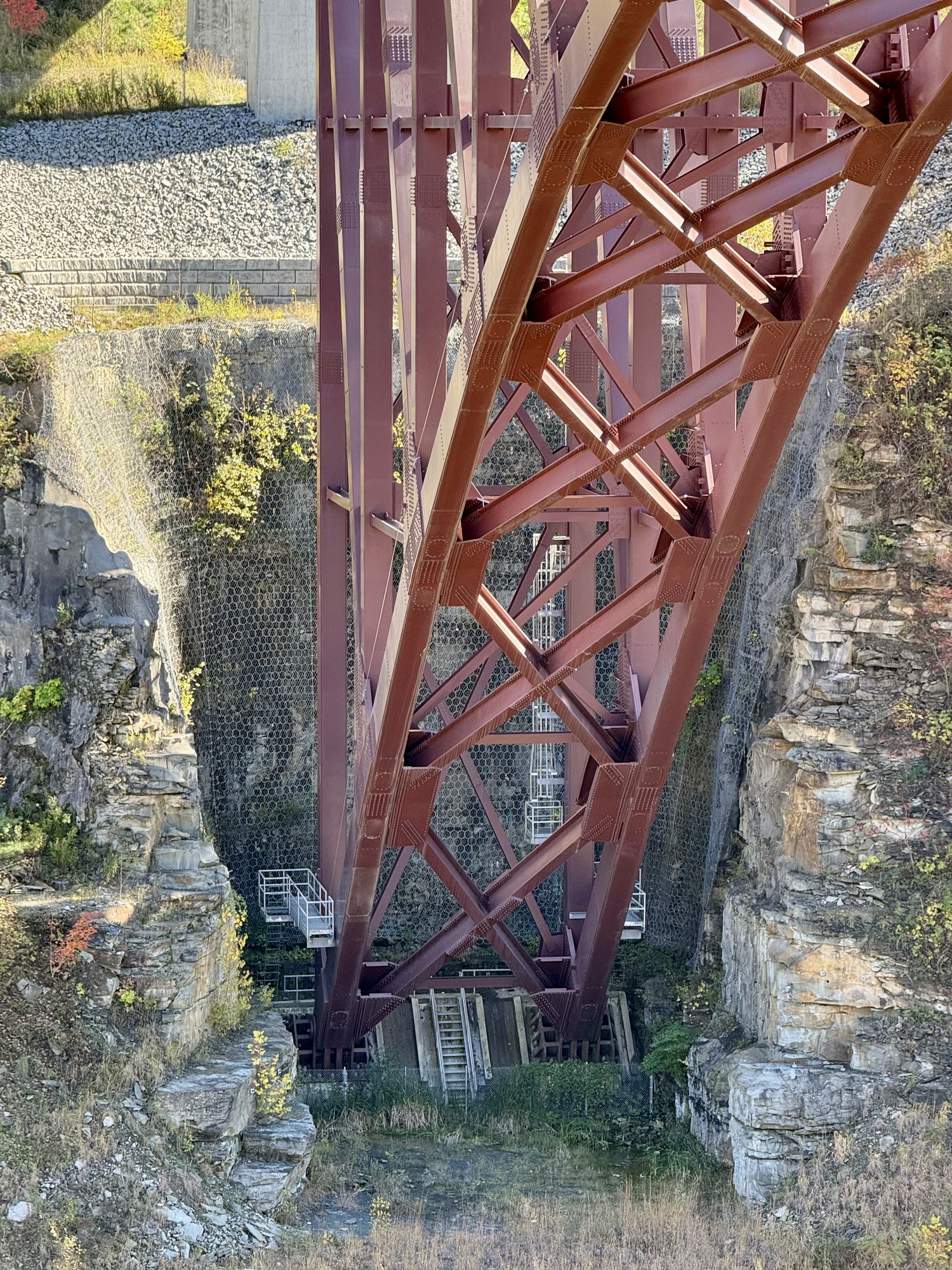 A telephoto shot of the eastern base of the Genesee Arch Bridge (also known as the Portage Viaduct). The red steel arch extends beyond the frame. The base is embedded into an area likely excavated out of the gorge’s wall. Various stairs and catwalks line the sides of the base.