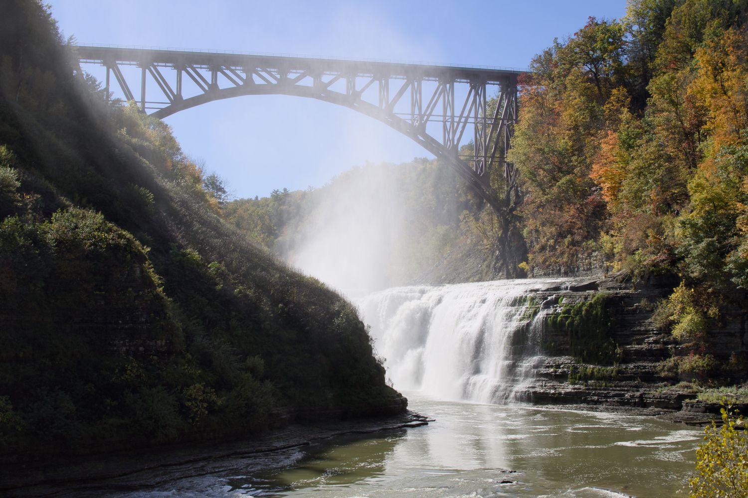 A waterfall in a gorge, surrounded by trees beginning to take on fall colors. The midday sun brightly lights the falls and the mist filling the gorge. A long train bridge spans the gorge over the falls.