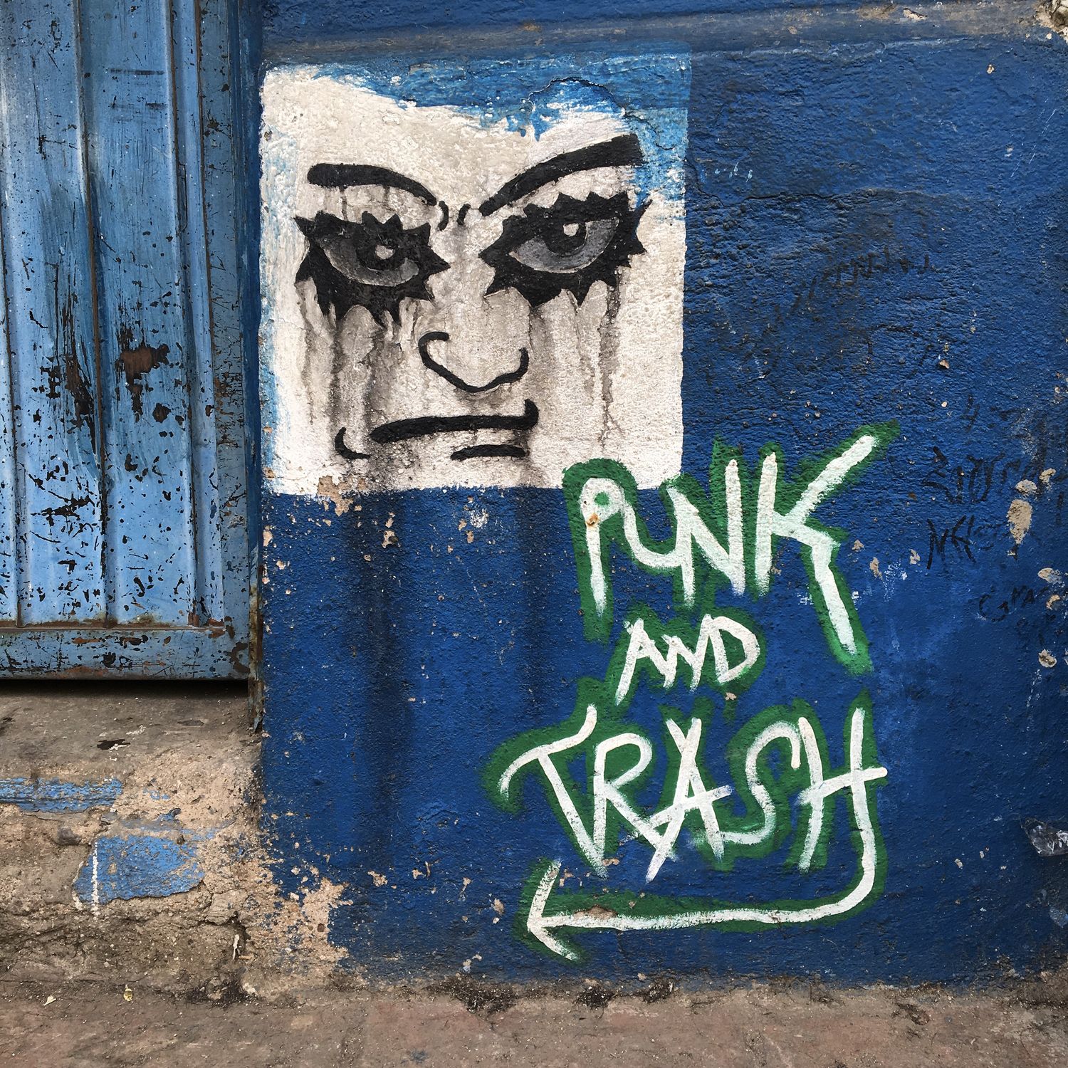 Close-up of a raw, rebellious piece of street art on a weathered blue wall. A black-and-white stencil of a square face with intense, smudged eyes stares out, its heavy eyeliner dripping like tears. Below it, graffiti in bold green and white reads “PUNK AND TRASH,” with an arrow pointing the way toward the entryway of the building — a worn blue metal door.